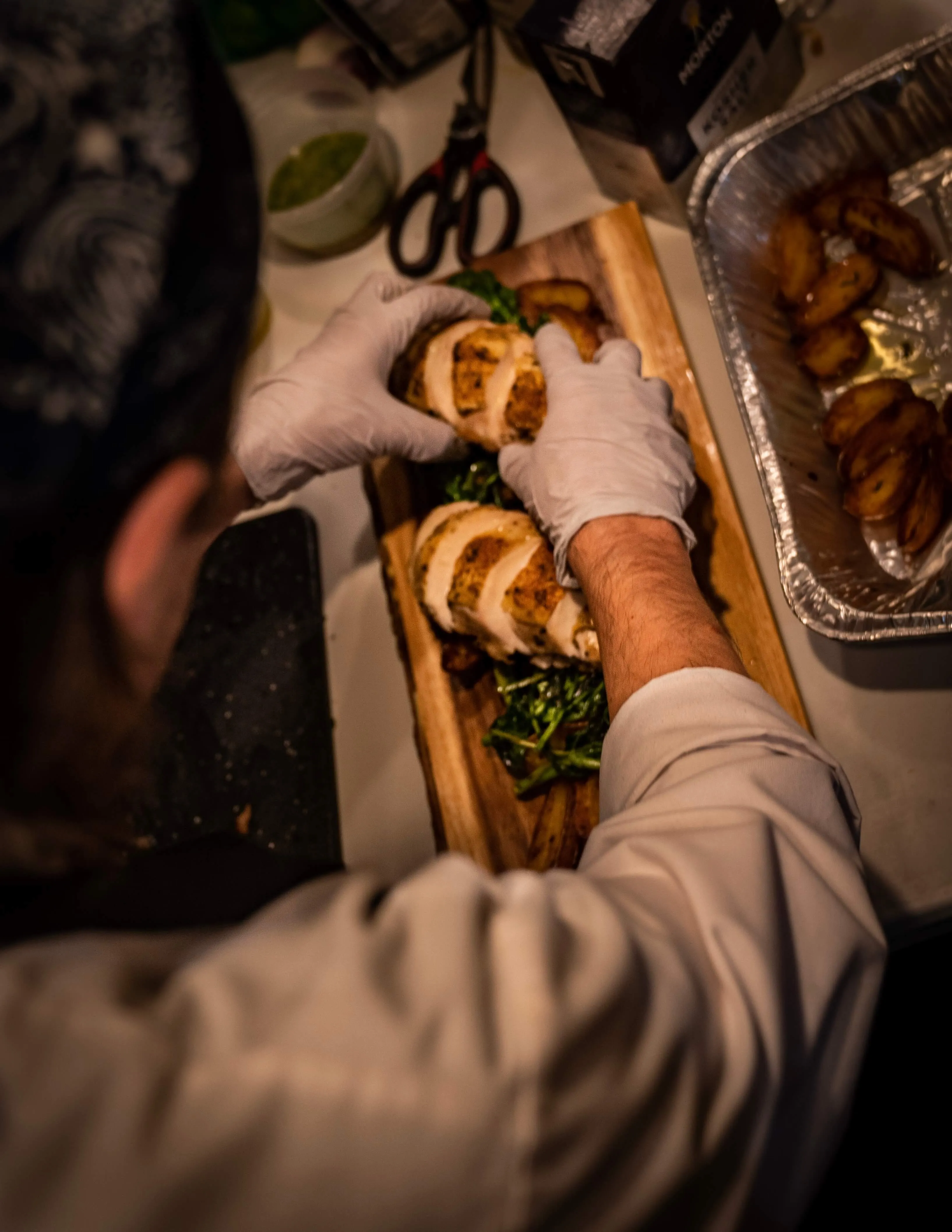 Chef in gloves plating sliced chicken and greens on a wooden board